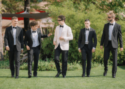 Elegant wedding party of friends in tuxedos enjoying a sunny outdoor celebration.